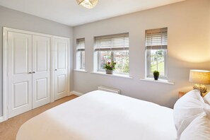 A bedroom with a large bed, a wardrobe, two windows with blinds, a radiator, and a bedside lamp. - Host Stay The Tardis (Redcar)