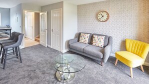A living room with a grey sofa, yellow armchair, and a glass coffee table. There is a clock on the wall and a door leading to another room.
