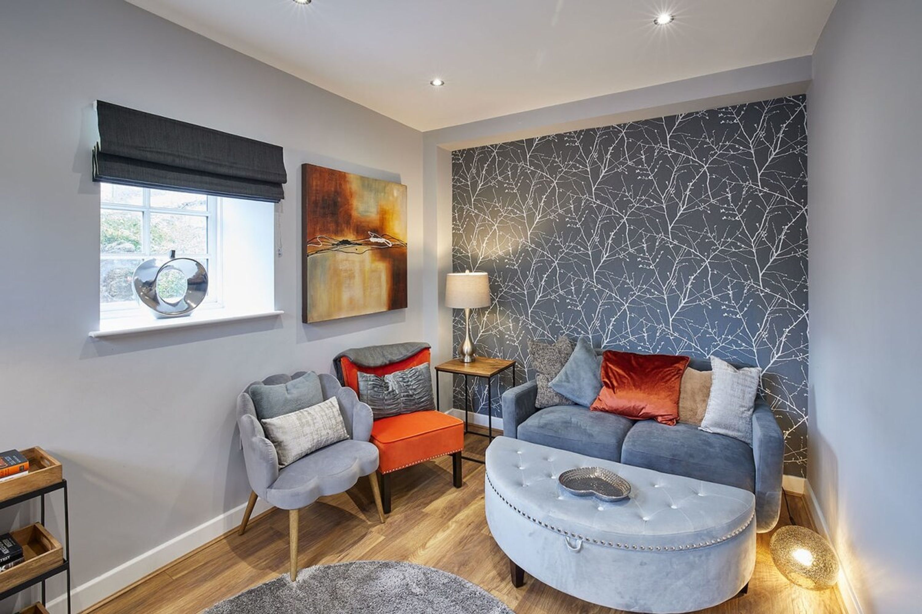 A modern living room with a grey sofa, a round ottoman, and a small side table with a lamp.
