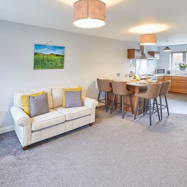 A modern living room with a white sofa, a dining table with four chairs, and a kitchen in the background.