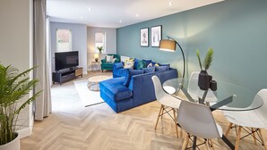 A modern living room with a blue sofa, a glass dining table, and a TV in the background. - Host Stay Turnstones Apartment (Whitby)