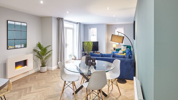 A modern living room with a glass dining table, white chairs, a blue sofa, a flat-screen TV, and a fireplace. - Host Stay Turnstones Apartment (Whitby)