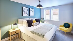 A bedroom with a bed, bedside table, lamp, framed botanical prints, windows, and a yellow armchair. - Host Stay Turnstones Apartment (Whitby)