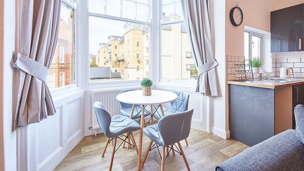A cozy dining area with a round table, four chairs, and a view of buildings through large windows. - Host Stay The Lookout (Scarborough)