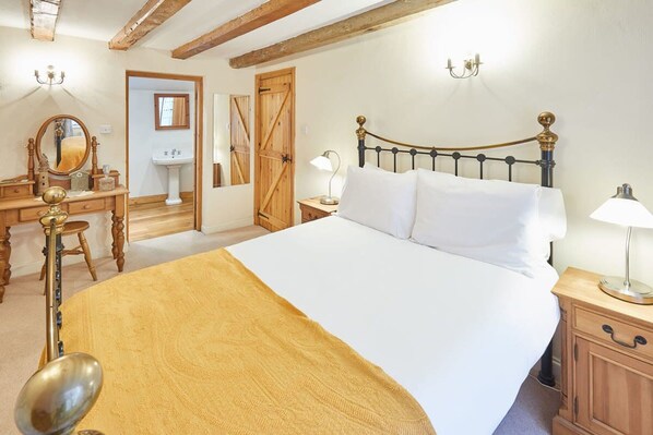 A bedroom with a large bed, wooden headboard, bedside tables with lamps, a dresser, a mirror, and a door leading to a bathroom. - Host Stay Jacksons Cottage (Whitby)