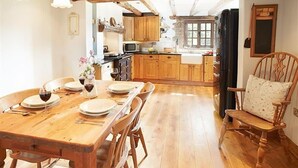 Ein Holztisch ist gedeckt mit Tellern und Weingläsern, ein Holzstuhl steht daneben. Im Hintergrund eine rustikale Küche mit Holzschränken und einem Fenster.