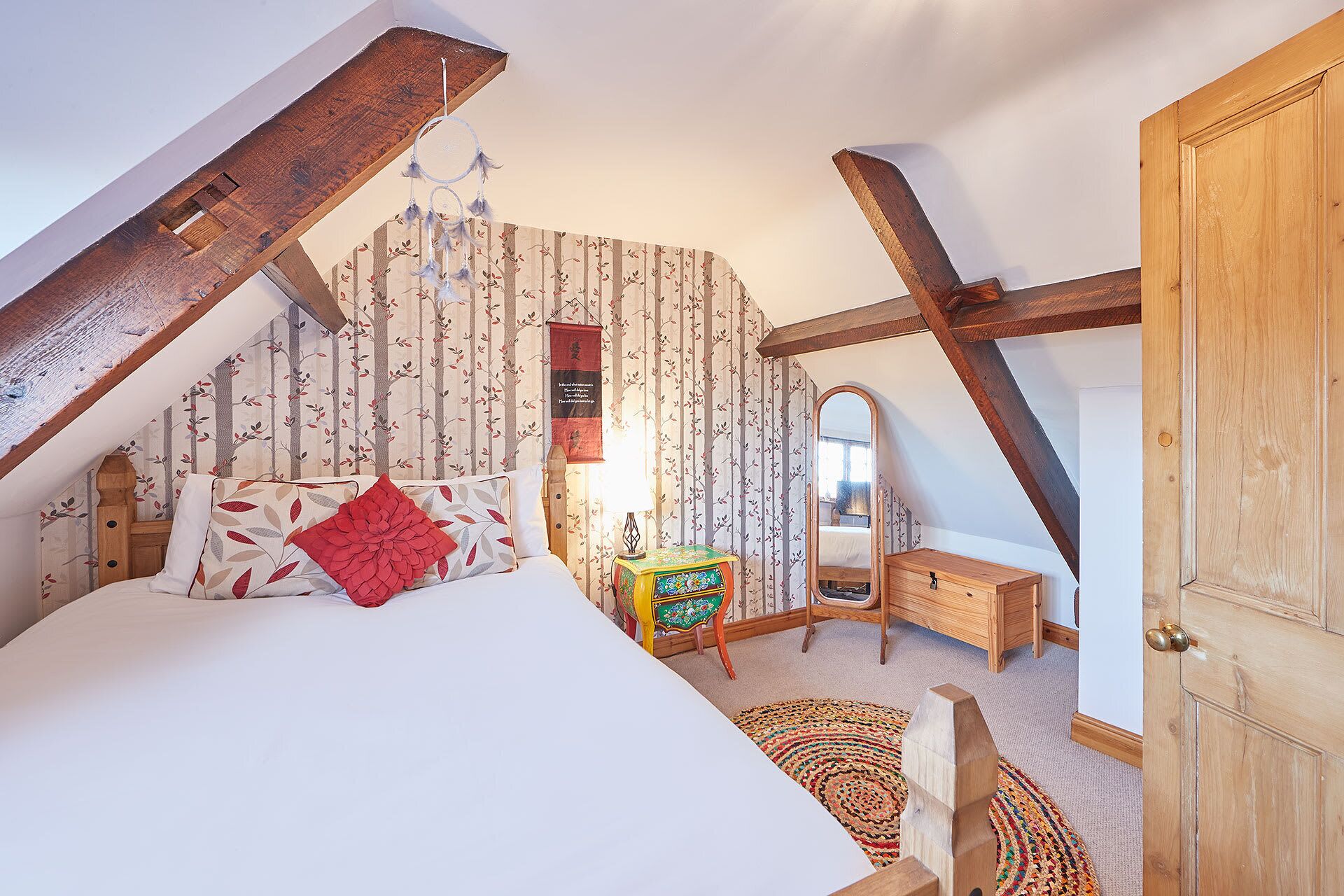 A bedroom with a sloped ceiling, a bed with a red pillow, a small wooden nightstand, a mirror, and a door.