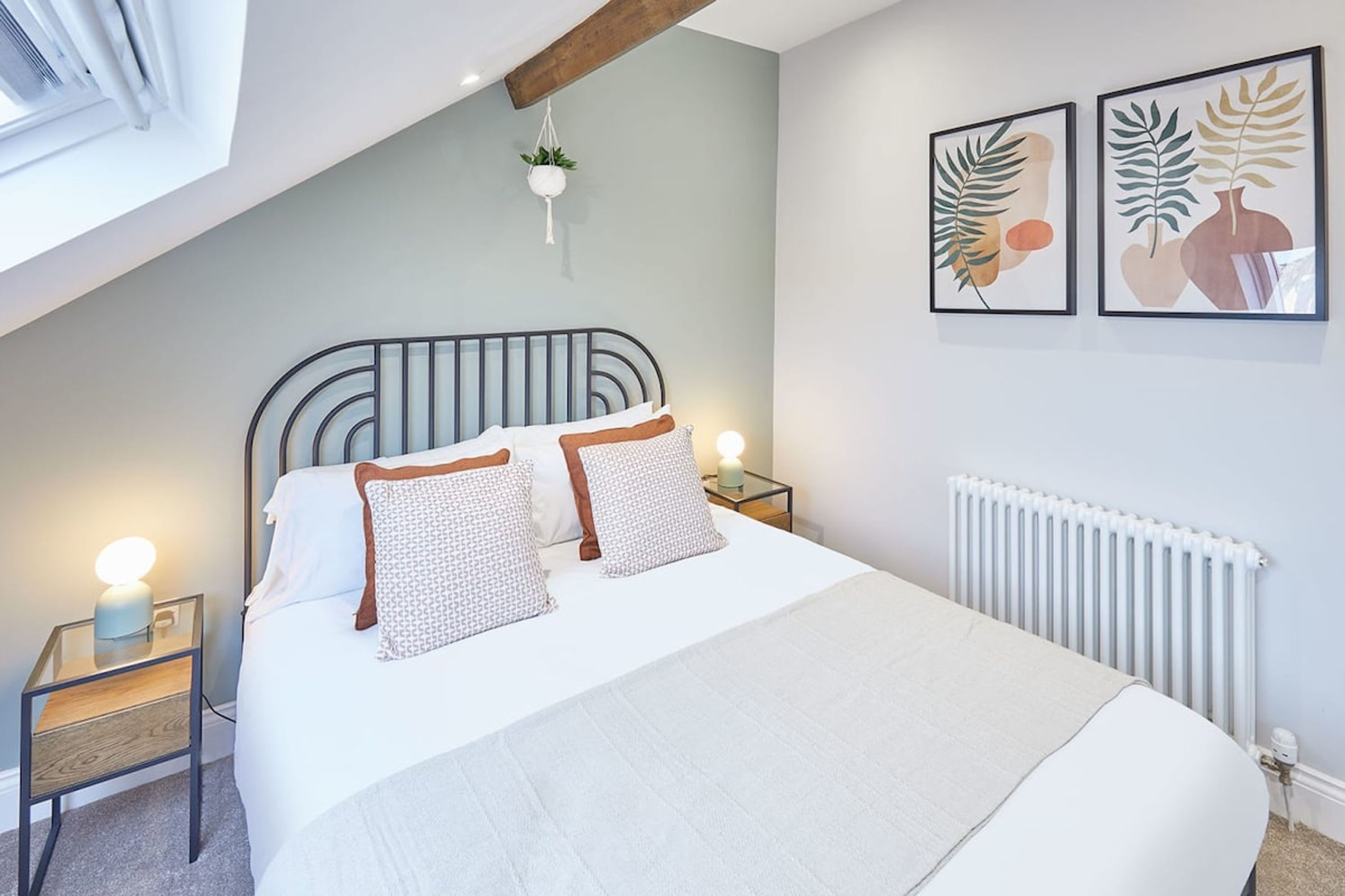 Une chambre à coucher avec un lit, des tables de chevet, une plante suspendue et des œuvres d’art encadrées accrochées au mur.