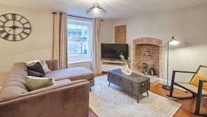 A living room with a sofa, a coffee table, a television, a brick fireplace, and a clock on the wall. - Host Stay Sleepers No 8 (Pickering)