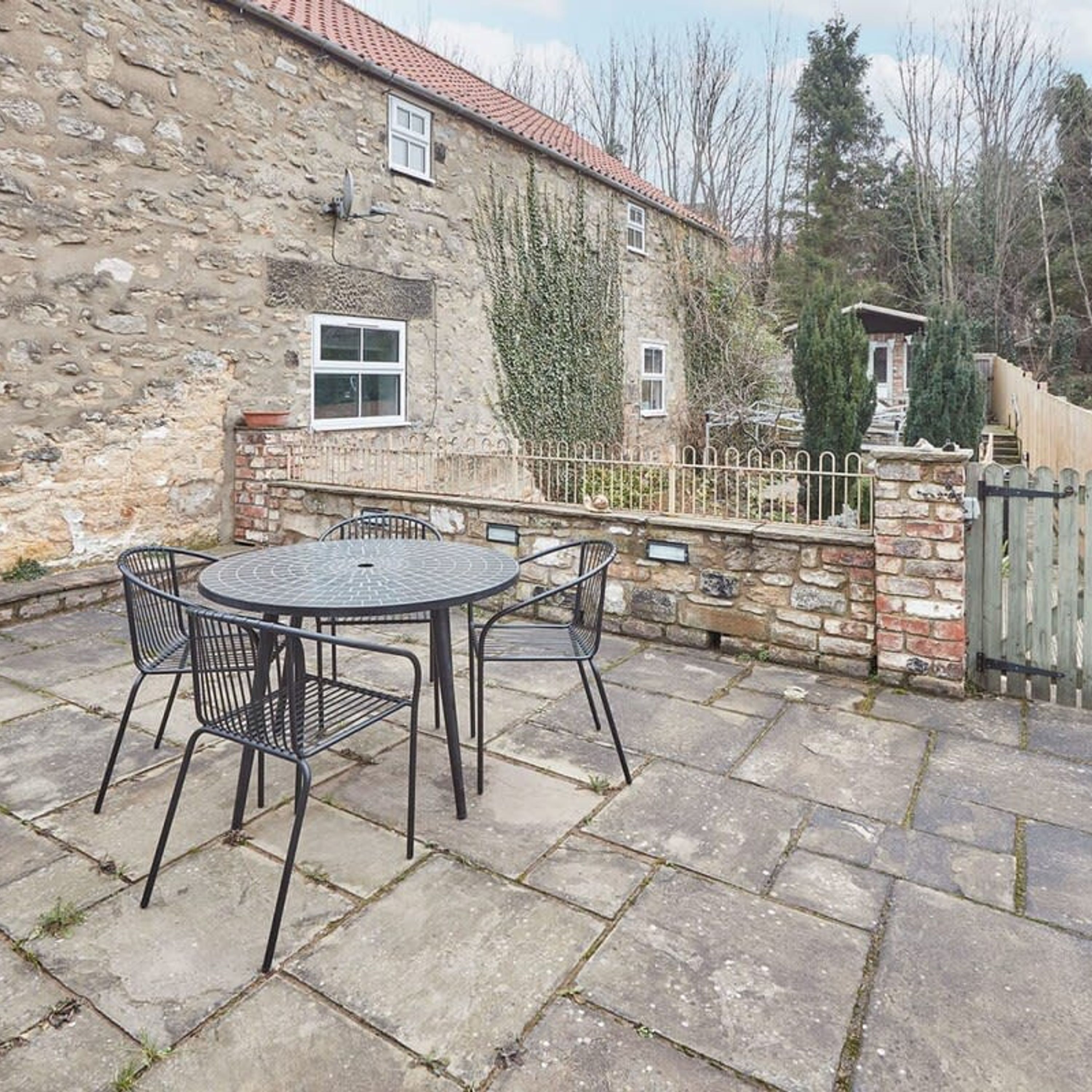 Ein Steinhaus mit Ziegeldach, einem umzäunten Garten und einem runden Tisch mit Stühlen.