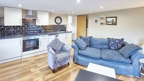A modern kitchen with white cabinets, a built-in oven, and a black countertop. There is a grey armchair and a blue sofa with patterned cushions. - Host Stay Abbey Heights (Whitby)