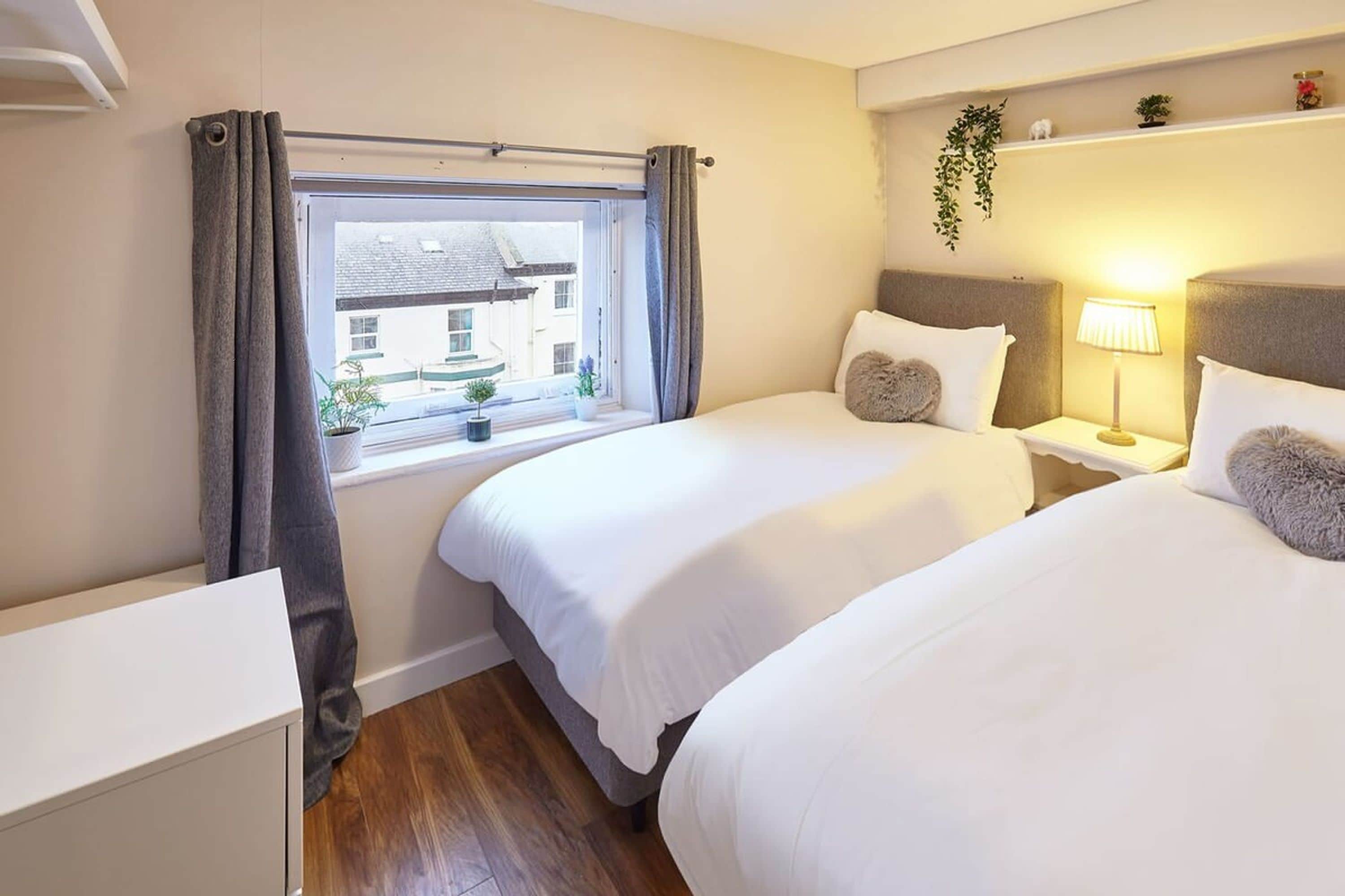 A hotel room with two beds, a window with curtains, a bedside table with a lamp, and a small plant on the windowsill.