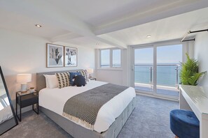 A modern bedroom with a large bed, a nightstand with a lamp, a blue chair, and a balcony with a view of the sea. - Host Stay Penthouse Pier View (Whitby)
