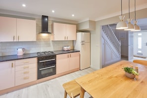 A modern kitchen with a gas stove, oven, and refrigerator, wooden dining table, and a staircase leading to another level.