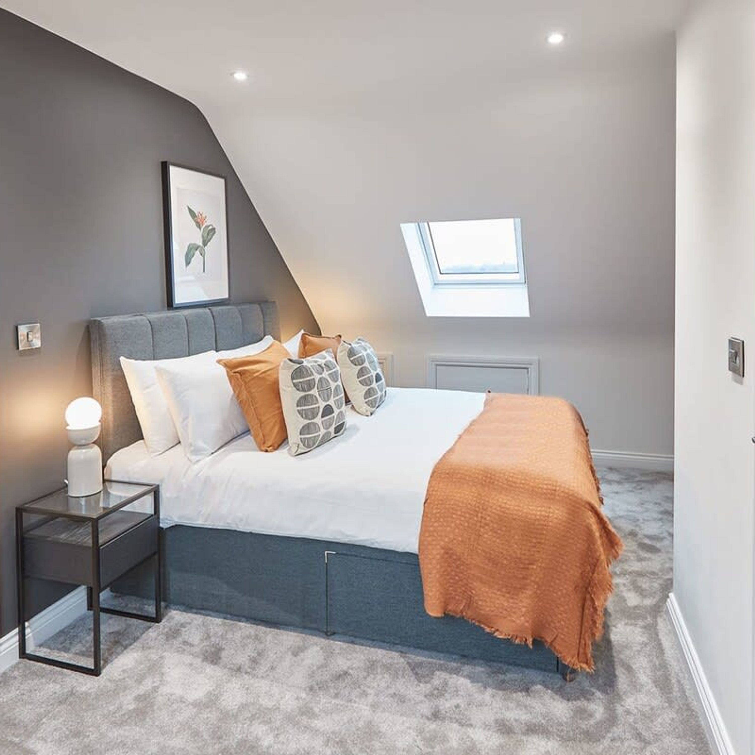 A bedroom with a sloped ceiling, a skylight, a bed with a grey headboard, and a bedside table with a lamp.