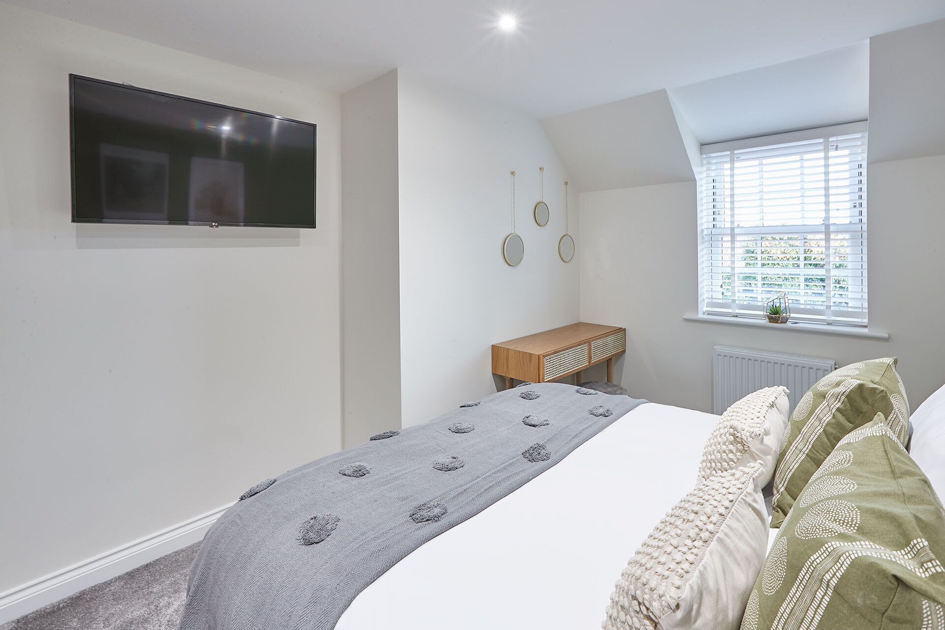 Une chambre à coucher avec un lit, un téléviseur fixé au mur, une petite table en bois et une fenêtre avec des stores.