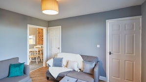 Una sala de estar con un sofá gris, un reposapiés gris y un cojín azul. Se observa una puerta blanca y un marco de madera.