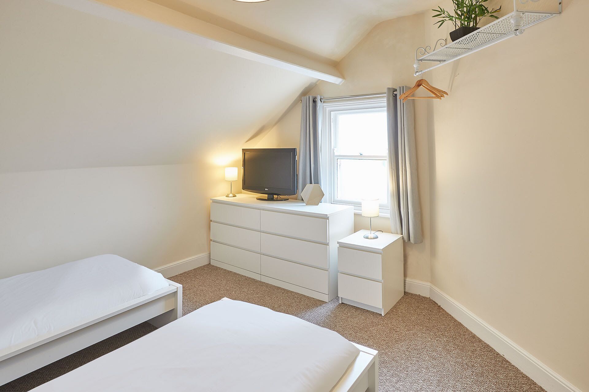 A small, well-lit bedroom with a single bed, a desk with a computer and lamp, a TV, and a window with curtains.