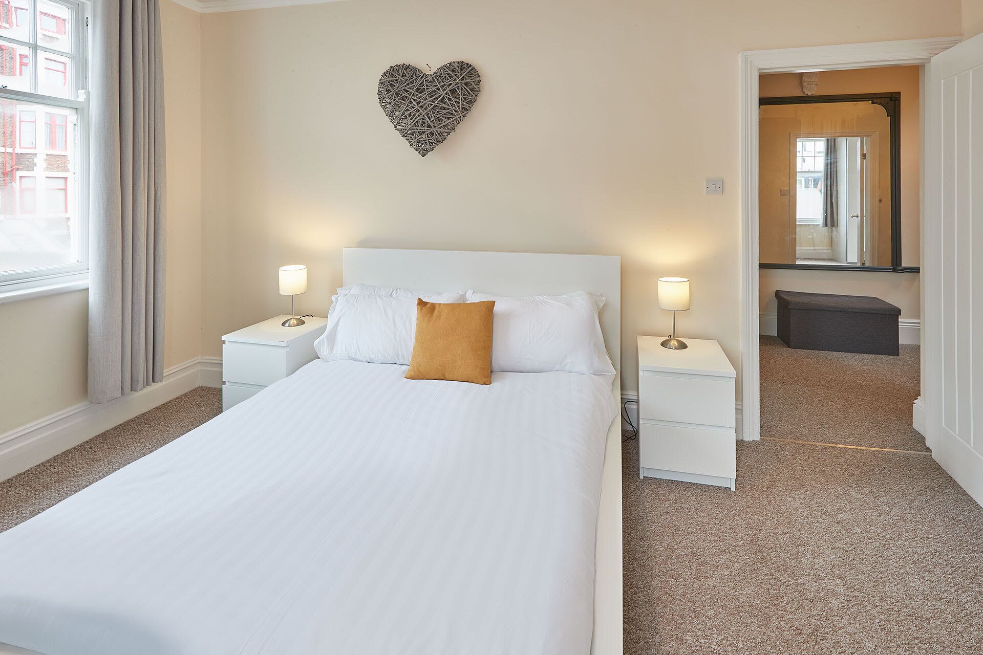 Une chambre d’hôtel bien aménagée, avec un grand lit, deux tables de chevet avec des lampes, une décoration murale en forme de cœur et un banc placé dans un coin.