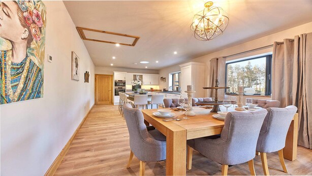 A modern dining area with a wooden table and grey upholstered chairs, a chandelier, and a view of the outdoors through a large window. - Host Stay The Parlour (Newcastle-upon-Tyne)