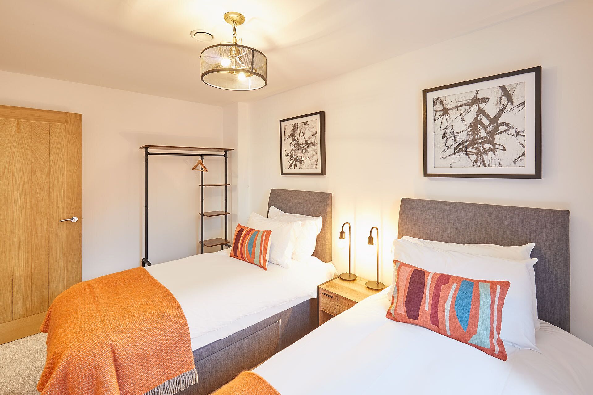 A hotel room with two beds, a wooden door, a metal rack, and abstract art on the walls.