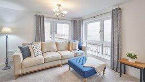 A living room with a beige sofa, a blue ottoman, a floor lamp, and a view of the sea through large windows.
