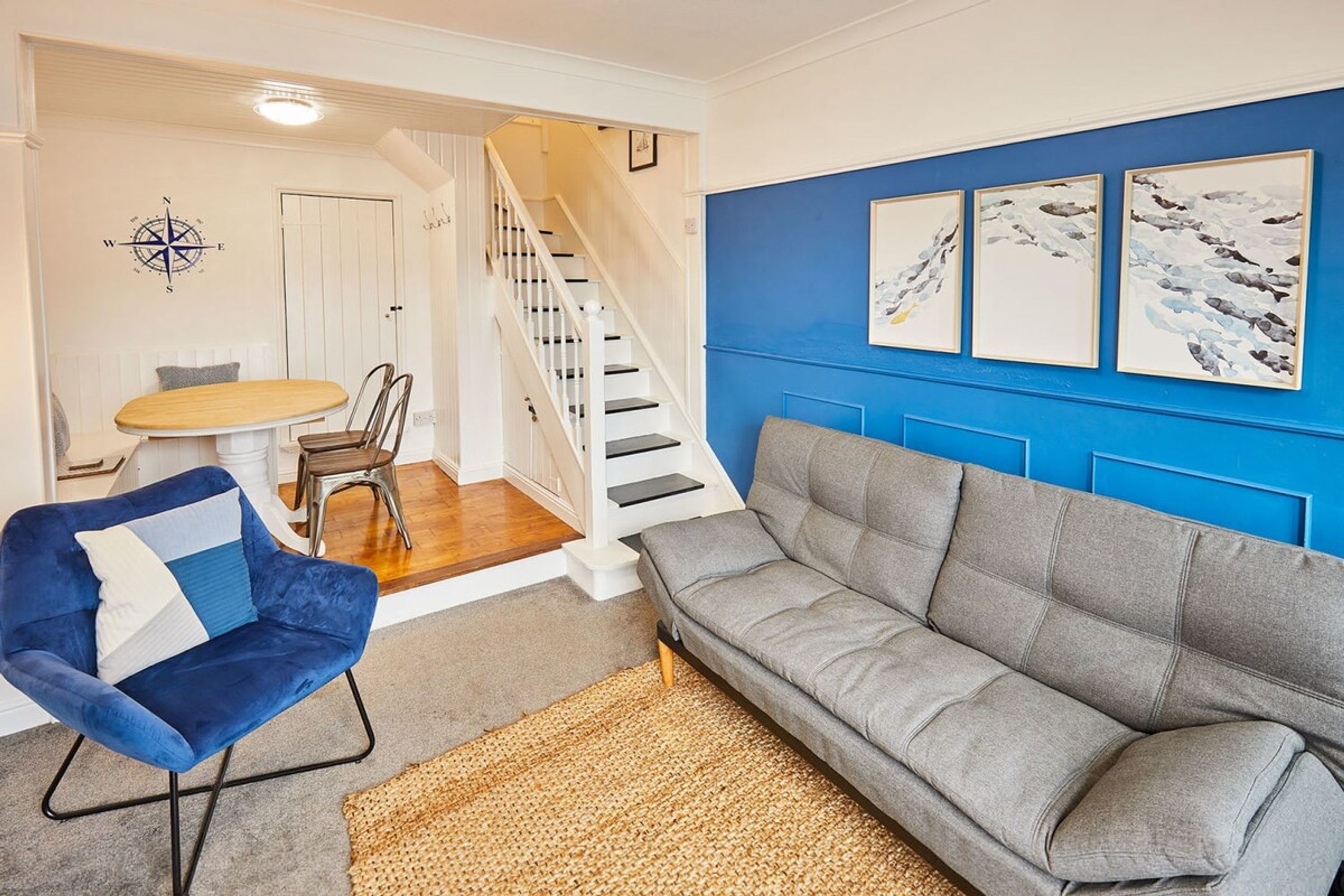 Een woonkamer met een blauwe accentmuur, een grijze bank, een blauwe fauteuil, een houten eettafel en een trap.