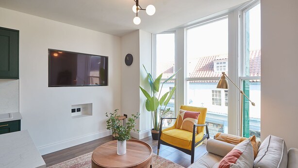 A modern living room with a sofa, a yellow armchair, a round wooden coffee table, a TV mounted on the wall, and a large window with a view of buildings. - Host Stay Abbey Nook (Whitby)