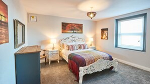 A bedroom with a white ornate bed, two bedside tables with lamps, a large window with blinds, and a painting of a tree on the wall. - Host Stay Seas The Day (Saltburn-by-the-Sea)