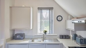 A modern kitchen with a built-in sink, a window with blinds, a clock on the wall, and a bread container labeled "BREAD" on the counter. - Host Stay Jet Cottage Spa Well (Whitby)