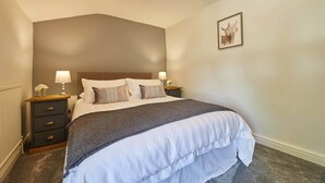 A bedroom with a large bed, two bedside tables with lamps, a framed picture on the wall, and a radiator under the window. - Host Stay Cummins Goings Cottage (Barnard Castle)