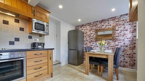 A compact kitchen with wooden cabinets, a microwave, a toaster, a refrigerator, and a dining area with a table and chairs. - Host Stay Cummins Goings Cottage (Barnard Castle)