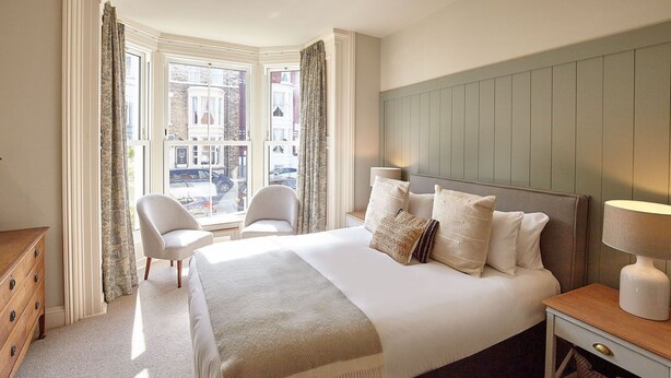 A hotel room with a large bed, a wooden dresser, a nightstand, and a view of the street through a bay window. - Host Stay The Salt Nook (Whitby)