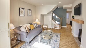 A modern living room with a grey sofa, a glass coffee table, and a patterned rug. There is a staircase leading to an upper level, and a TV mounted on the wall. - Host Stay The Salt Nook (Whitby)