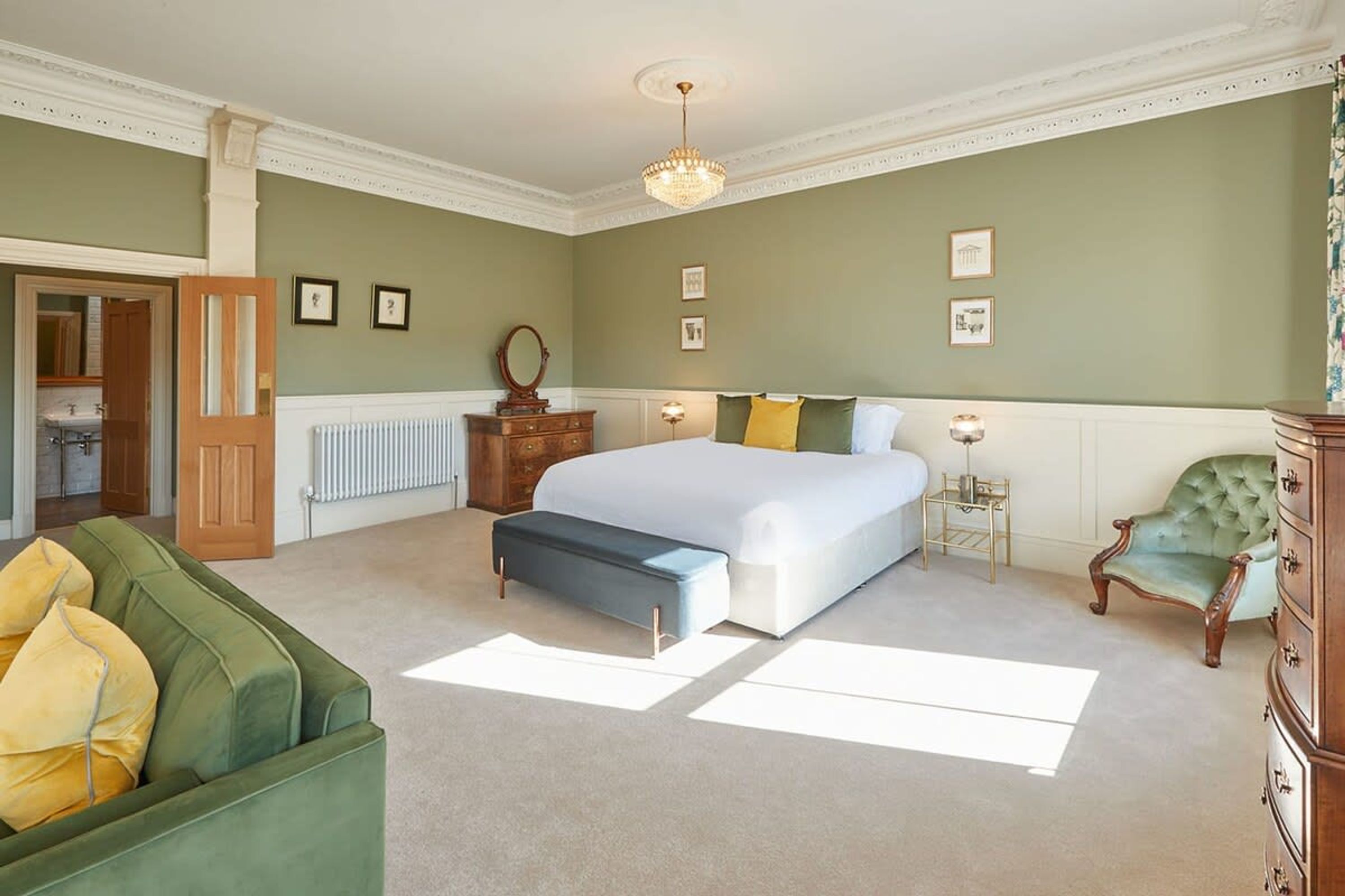 Een slaapkamer met een groot bed, een groene bank, een houten dressoir en een kroonluchter.