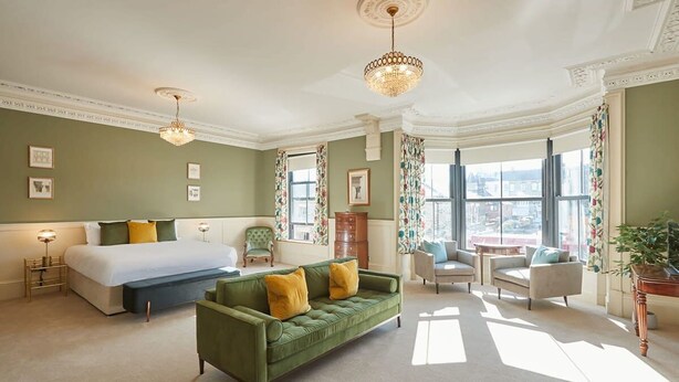 A spacious hotel room with a large bed, a green sofa, and a view of the city through large windows. - Host Stay Sandpiper (Saltburn-by-the-Sea)