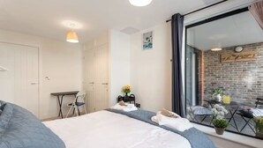 Quarto de hotel com cama, escrivaninha, cadeira e vista para uma varanda com plantas e parede de tijolos.