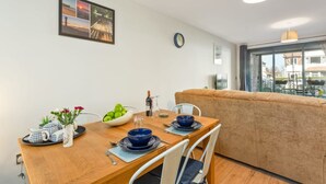 Uma sala de jantar com mesa de madeira posta para quatro pessoas, um sofá e uma grande janela com vista para edifícios.