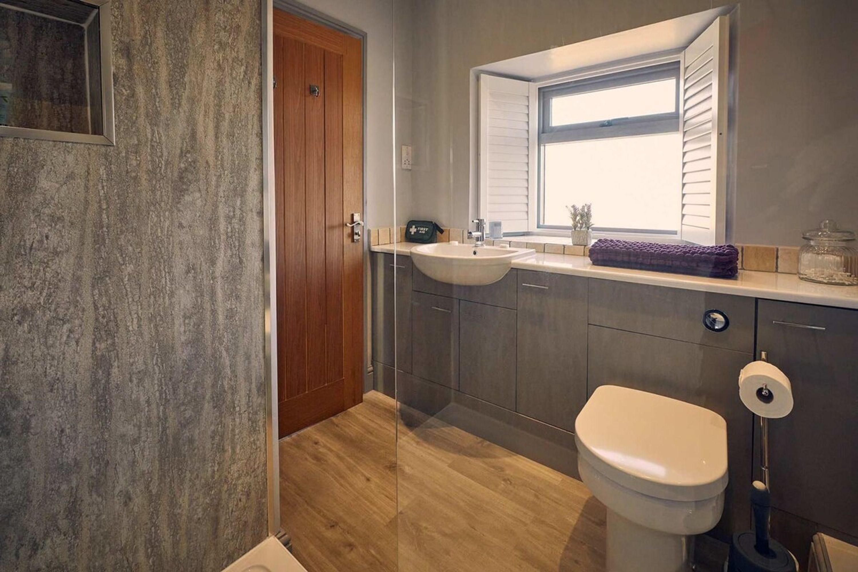 Un baño moderno con puerta de madera, un lavabo, un inodoro y una ventana con persianas.