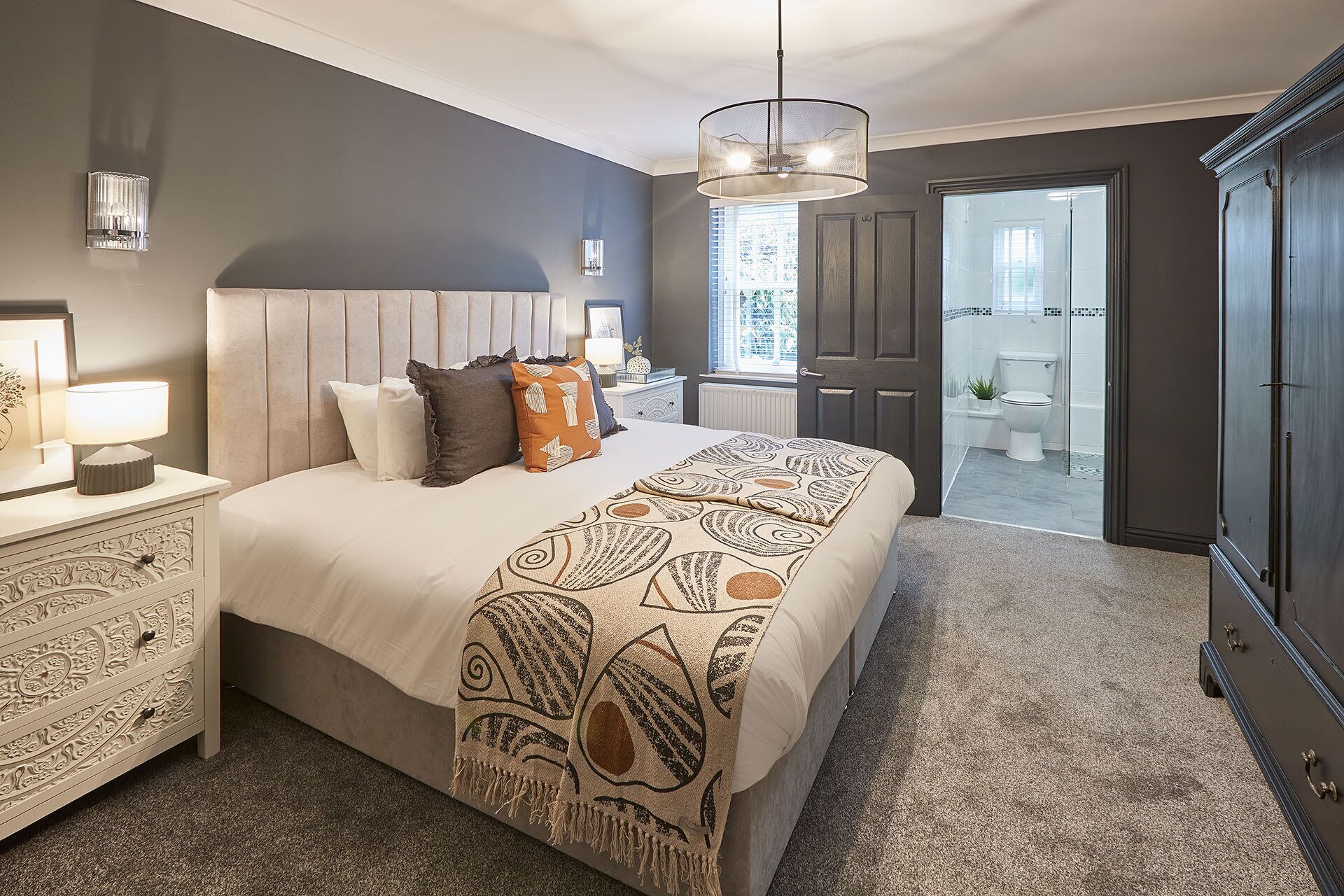 Een slaapkamer met een groot bed, een nachtkastje met lamp, een dressoir en uitzicht op de badkamer door een open deur.