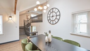 Un coin repas avec une table en bois foncé, des chaises recouvertes d’un tissu vert, deux vases blancs ornés de plantes, une grande horloge murale et un escalier menant à un autre niveau.