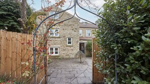 Une maison en pierre avec une cour dallée, une clôture en bois et un portail.