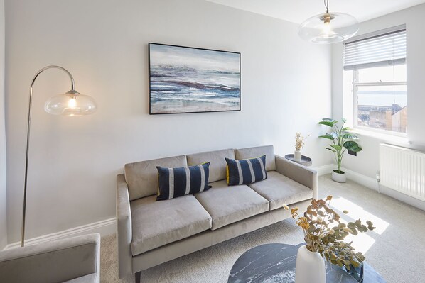 A modern living room with a grey sofa, a framed abstract painting, a floor lamp, a potted plant, and a view of the city through a window.