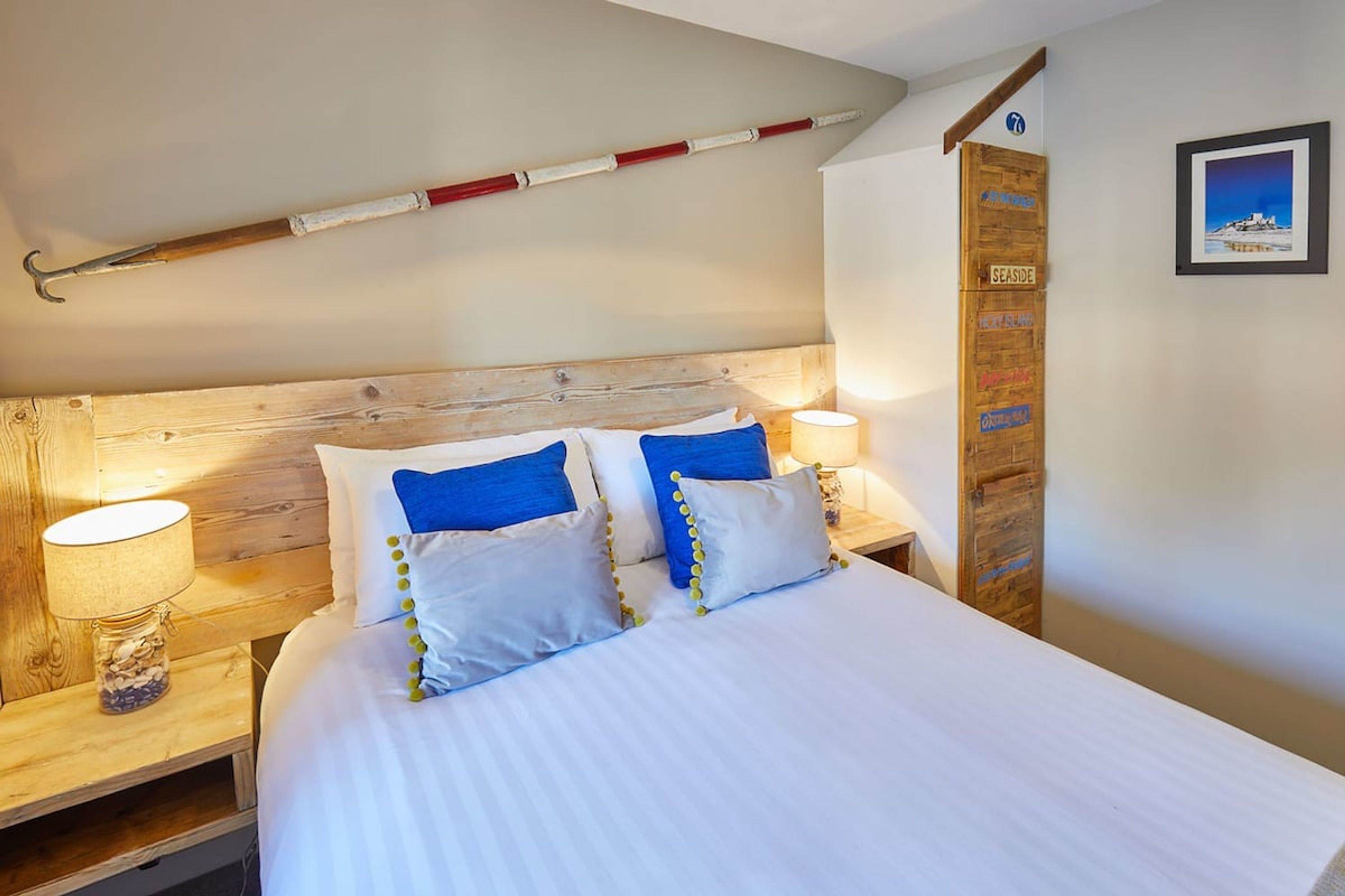 Un lit bien fait, avec une literie blanche et bleue, deux lampes de chevet, une tête de lit en bois, une échelle en bois et un mur orné d’une enseigne et d’un tableau encadré.