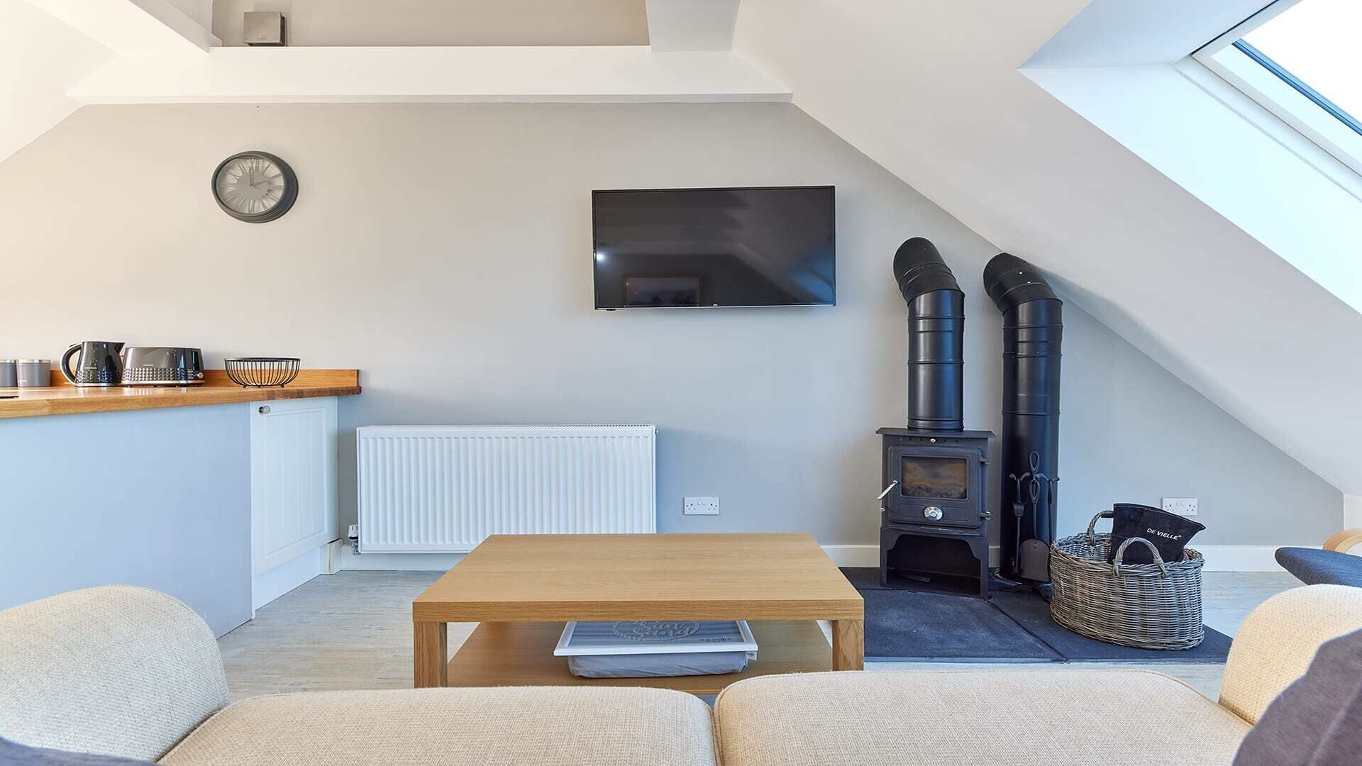 A cozy loft-style living space with a wooden table, a sofa, a TV mounted on the wall, and a wood-burning stove.