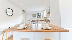 A modern kitchen with white cabinetry, a wooden island with two white bowls and wine glasses, and a window with a view of buildings. - Host Stay Ocean Pearl (Seahouses)