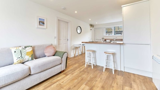 A modern living room with a sofa, two stools, a kitchen area with a fridge, and a door. - Host Stay Ocean Pearl (Seahouses)