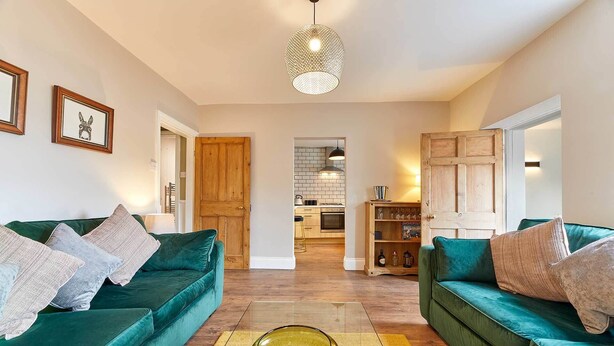 A modern living room with two teal sofas, a glass coffee table, and a kitchen visible through an open door. - Host Stay The Blacksmiths (Morpeth)
