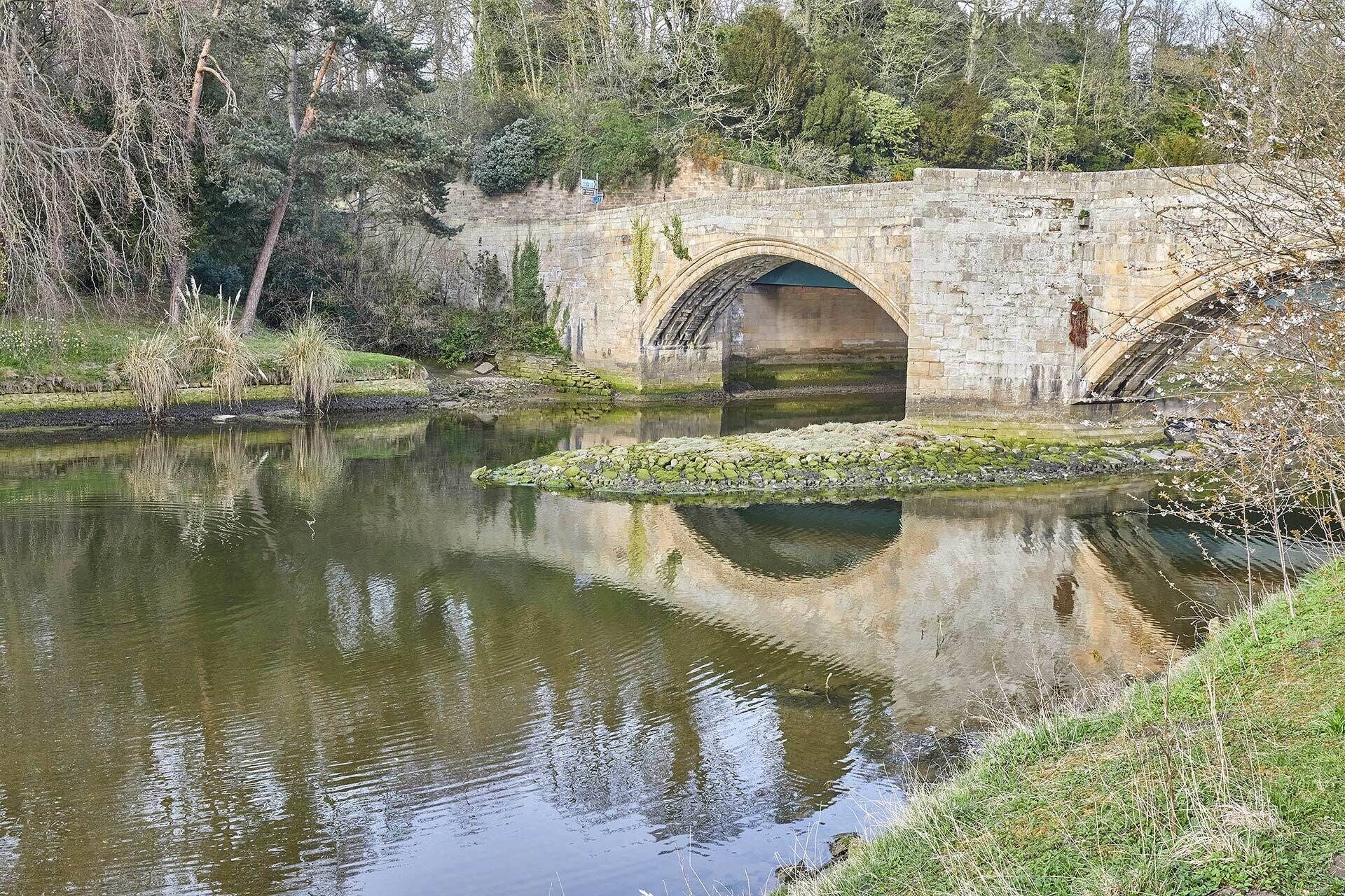 Un pont de pierre enjambant une rivière paisible, dont le reflet se mire dans l’eau, ainsi que celui des arbres qui l’entourent.