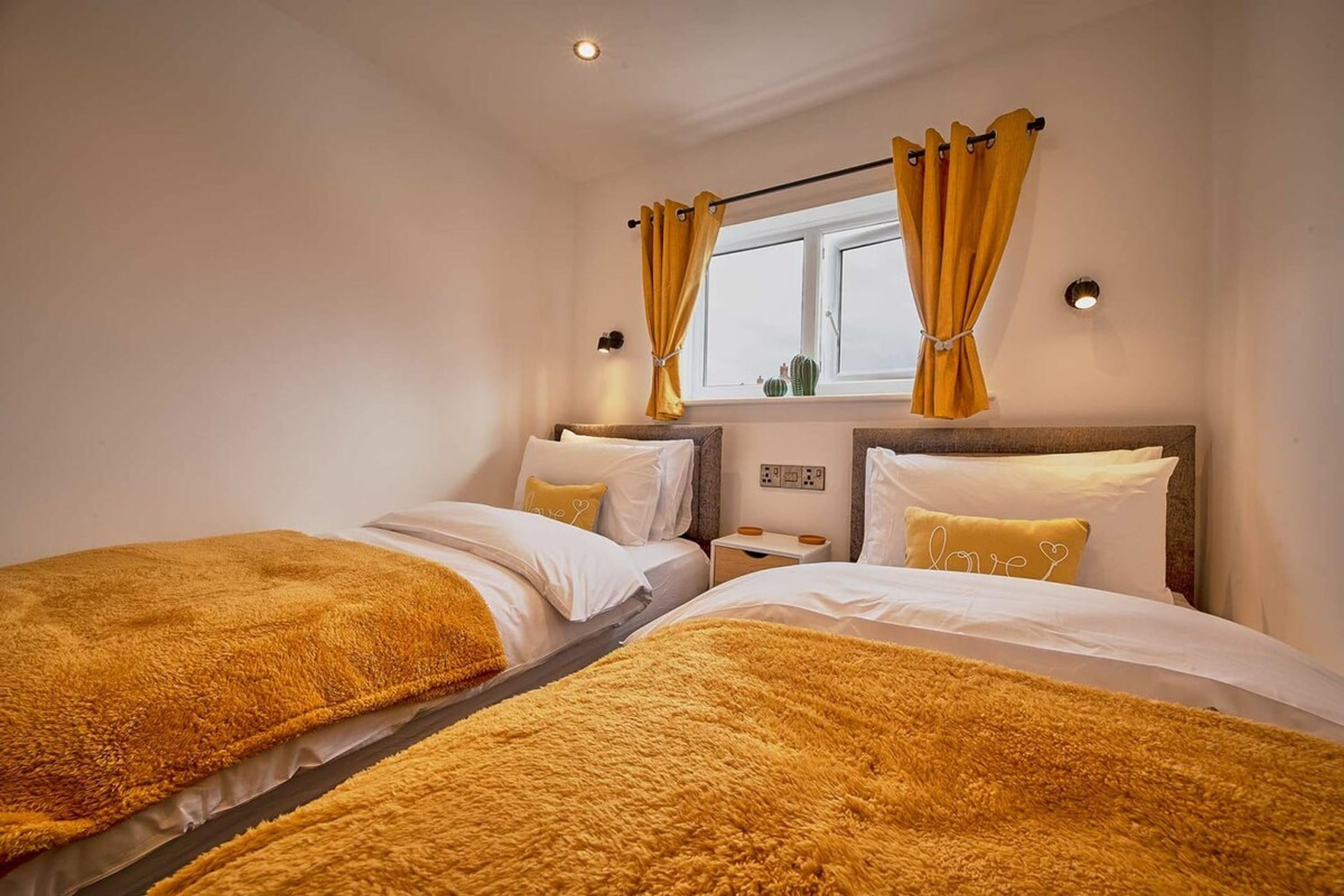 A hotel room with two beds, each with a fluffy golden blanket, white bedding, and a nightstand with a lamp.